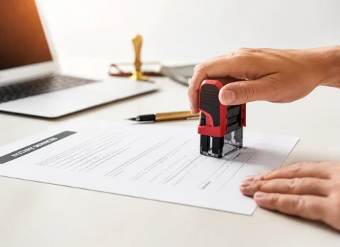 Hand using a notary stamp on a document beside a laptop and office workspace.
