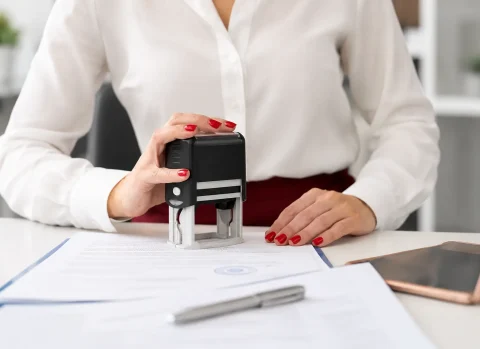 Professional office worker applying a notary stamp to a document at a desk with paperwork, pen, and mobile device nearby.